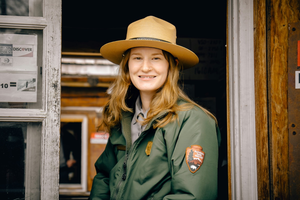 A park ranger in National Park Service uniform and flat hat, smiling in the doorway of a park entrance booth.